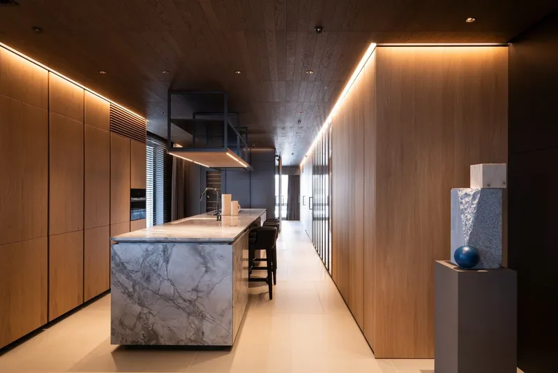 Long galley kitchen with marble island and continuous oak paneling under recessed ceiling lights