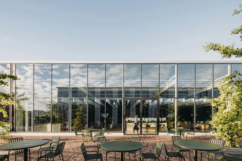 Outdoor brick terrace with green metal tables and chairs facing a floor-to-ceiling glass facade