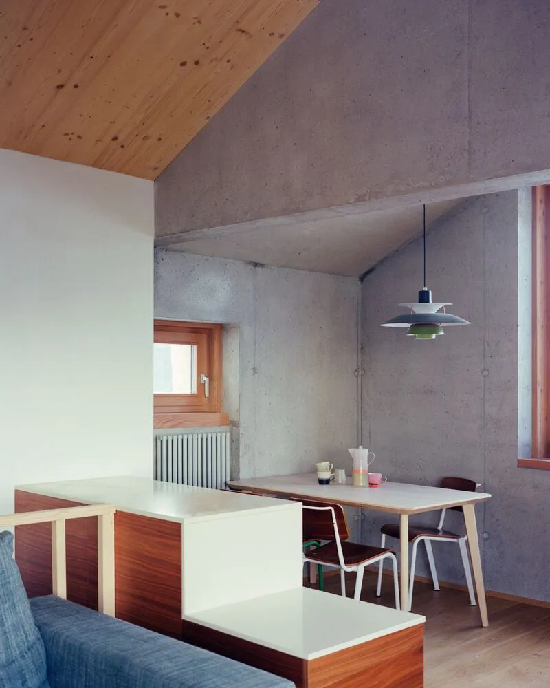 Interior showing exposed concrete walls and vaulted plywood ceiling above a dining area with pendant lamp