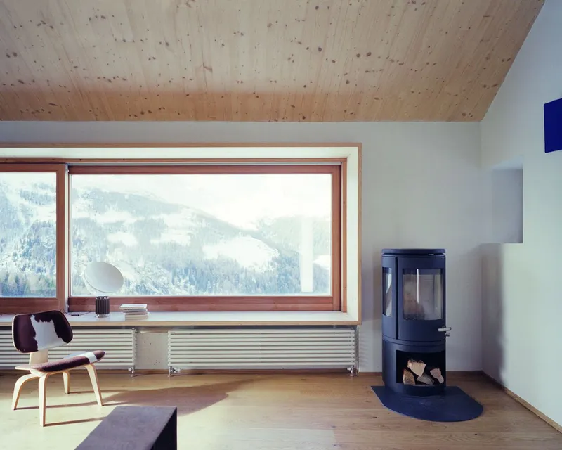 Wide timber-framed window with radiator below and freestanding wood stove overlooking snowy slopes