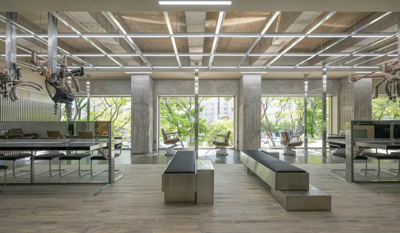 Salon interior with exposed concrete ceiling, steel grid structure, and stainless benches overlooking trees through full-height glazing