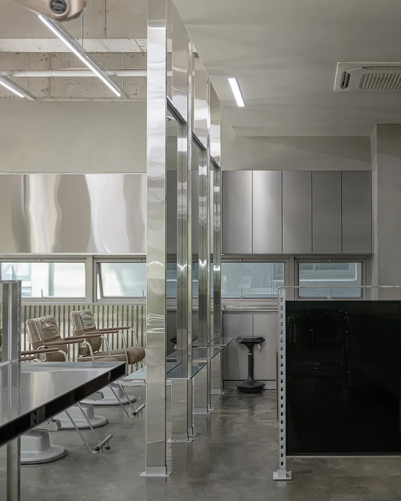 Reflective stainless steel column wrapping in salon space with industrial shelving and mirrored surfaces under linear lighting