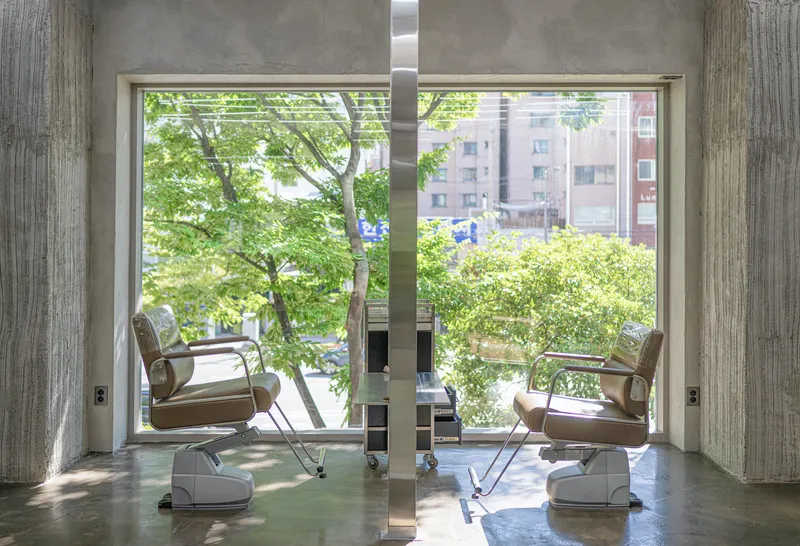 Framed window view between board-formed concrete columns showing salon chairs and green foliage beyond the glass