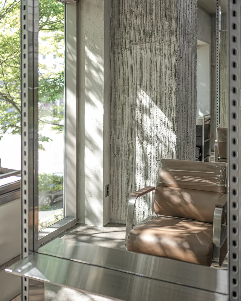 Detail of board-formed concrete column beside stainless steel shelving with dappled sunlight from adjacent trees