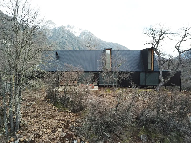 Linear metal-clad volumes on a rocky hillside with bare winter trees and snow-capped mountains beyond
