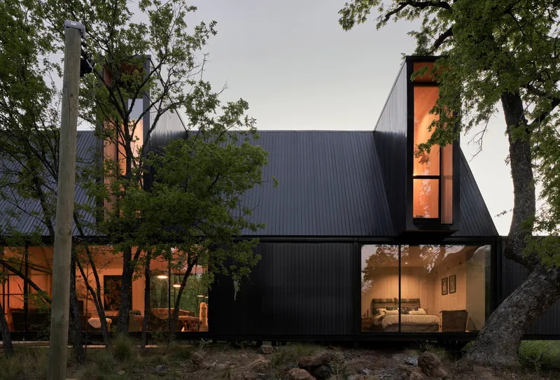 Dark metal-clad volumes with floor-to-ceiling glazing nestled among mature trees at twilight