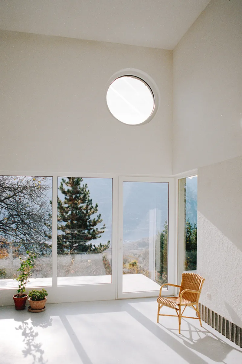 White interior room with circular ceiling window and floor-to-ceiling glazing overlooking hillside with wicker chair in sunlight