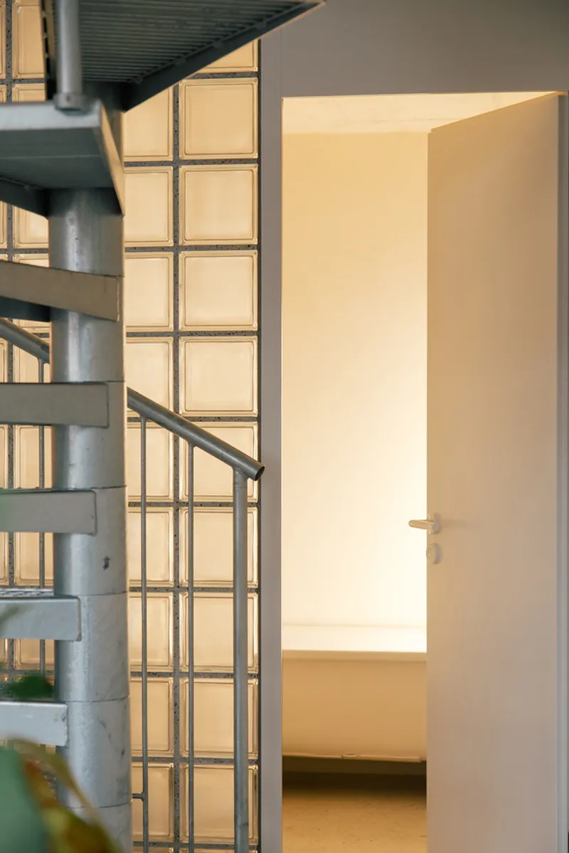 Metal spiral staircase beside translucent glass block wall with warm interior light glowing through