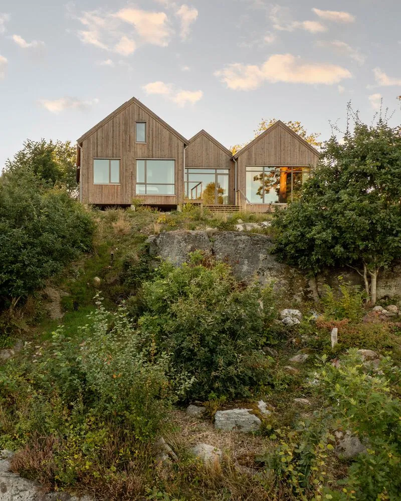 Three connected timber-clad gabled volumes elevated on columns above rocky outcrop at dusk