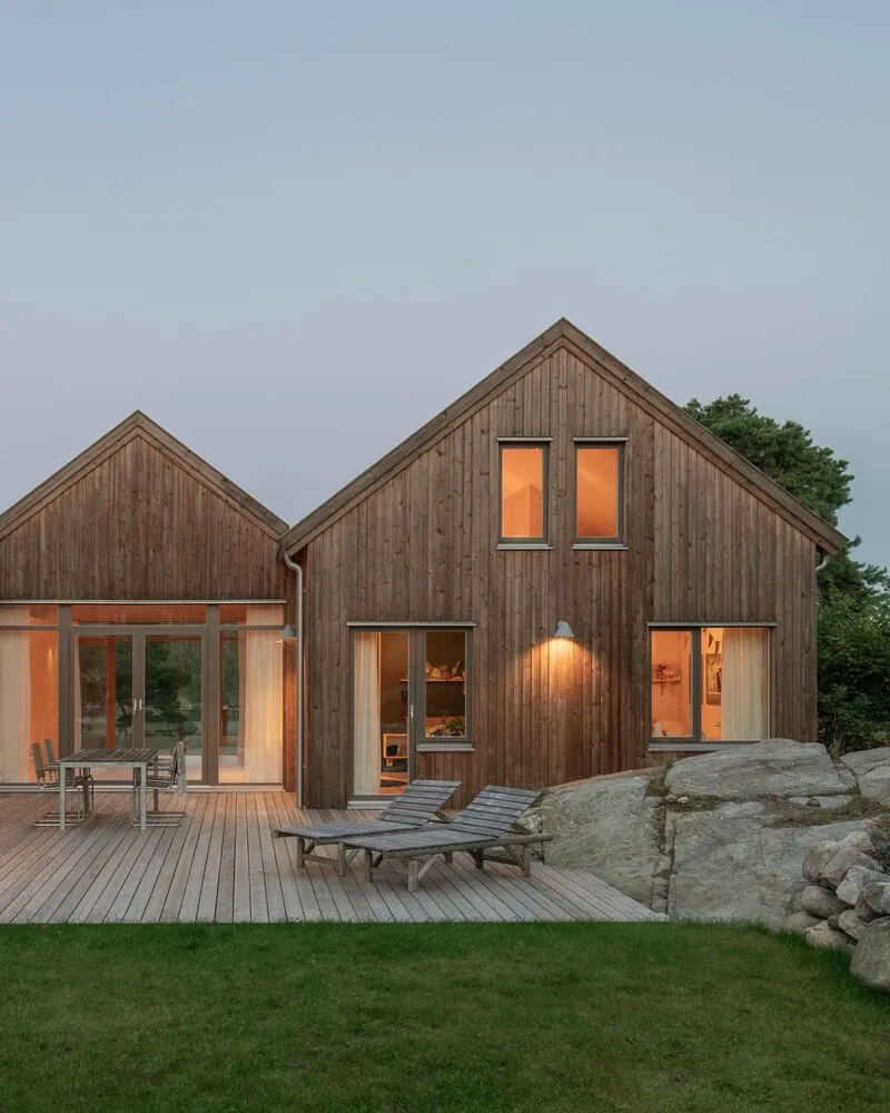 Timber deck with lounge chairs facing illuminated interior volumes at twilight beside granite outcrop