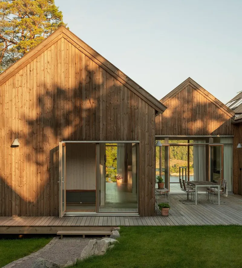 Vertical timber-clad gable facades with sliding glass doors opening onto a wooden deck with lawn and pine trees