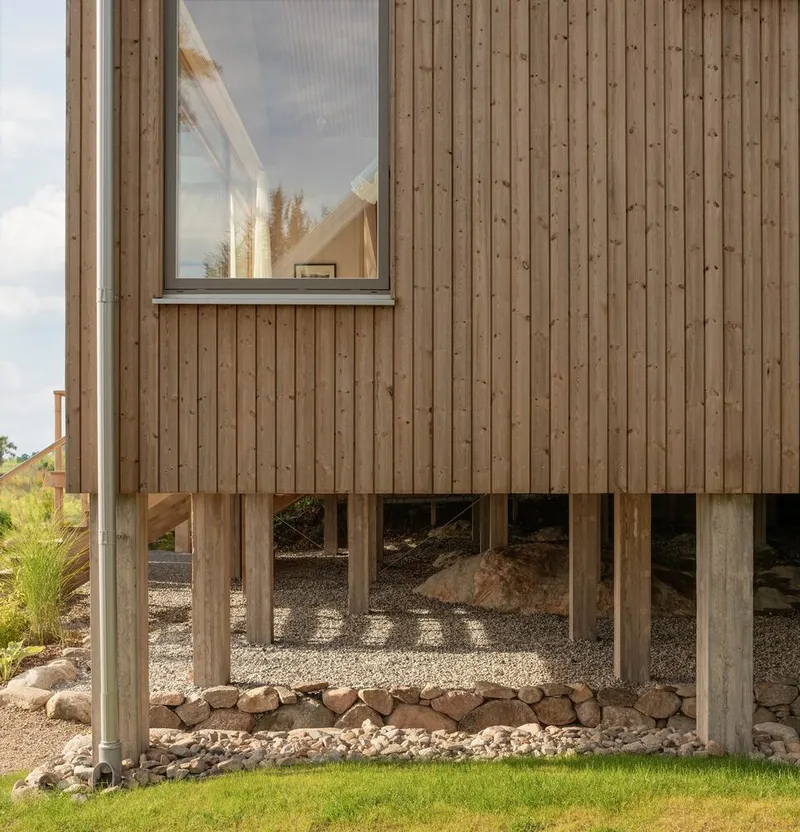 Timber-clad volume elevated on exposed wooden posts above a gravel bed with stone edging