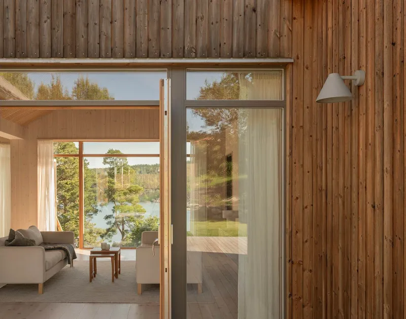 Vertical timber wall with glass doors opening to interior sitting room overlooking water through trees