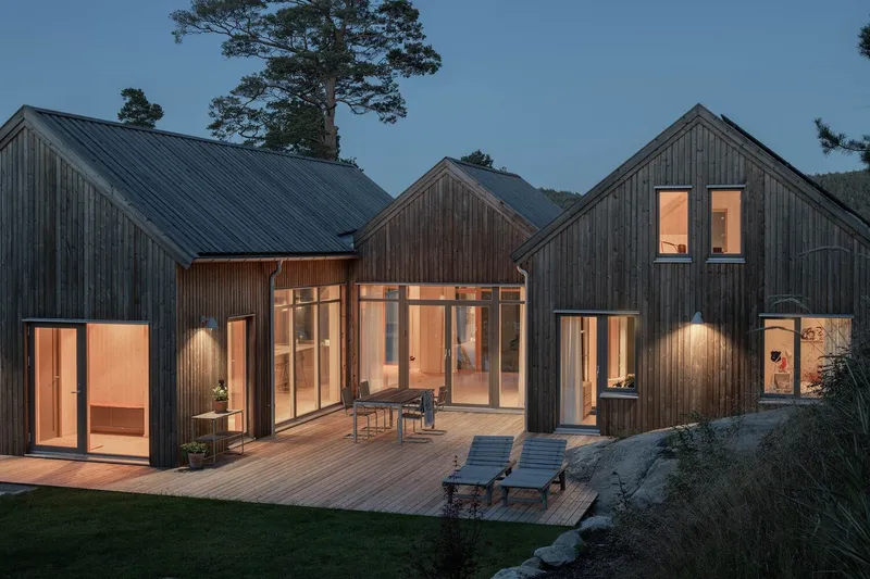 Cluster of timber-clad gable volumes with illuminated glazing surrounding a wooden deck terrace at twilight