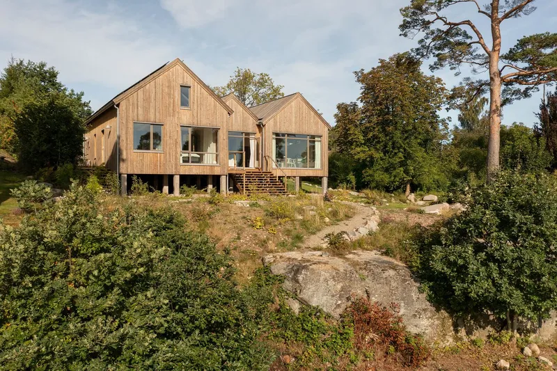 Timber-clad gabled volumes on pilotis above rocky terrain with scattered vegetation and pine trees