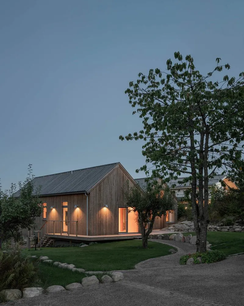 Timber-clad gable volume with illuminated wall sconces and glazed openings at dusk beside a deciduous tree