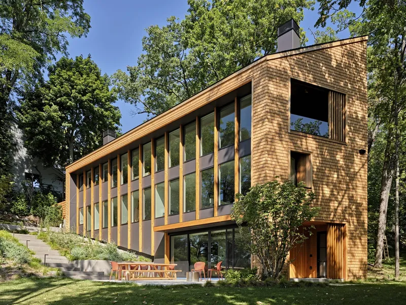 Cedar shingle facade with vertical timber and glass bays rising through a wooded site in afternoon sunlight