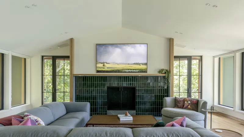 Living room with dark green tile fireplace surround flanked by floor-to-ceiling windows overlooking the trees