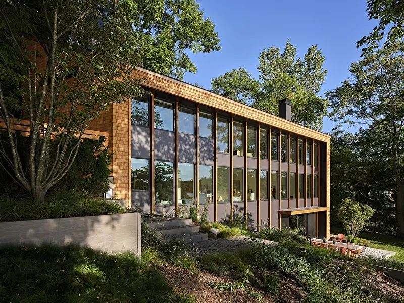 Cedar-clad volume with floor-to-ceiling glazing framed by deciduous trees on a sloped garden