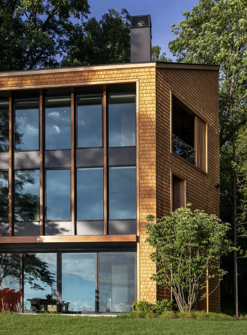 Cedar shingle facade with floor-to-ceiling glazing and metal chimney surrounded by mature trees at dusk