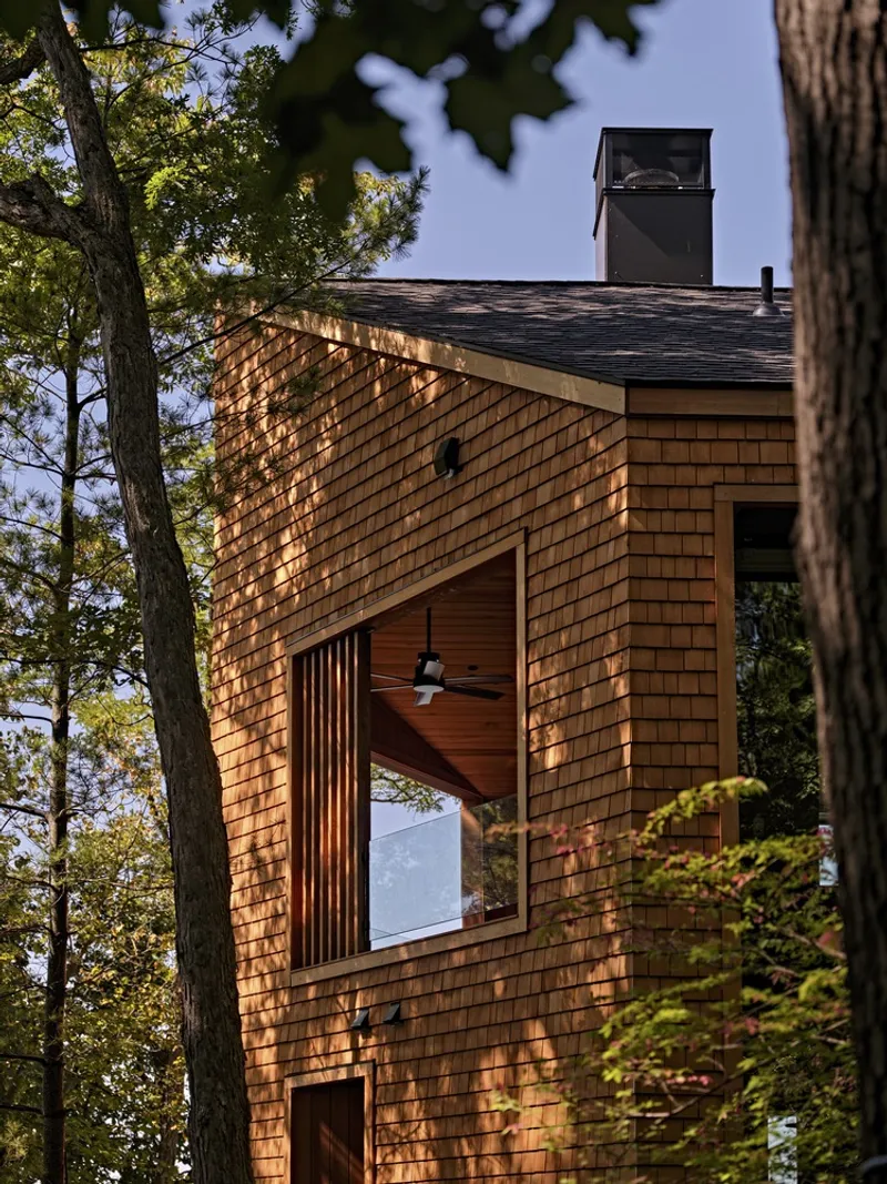 Wood-clad volume with vertical slot window framing a ceiling fan viewed through forest canopy