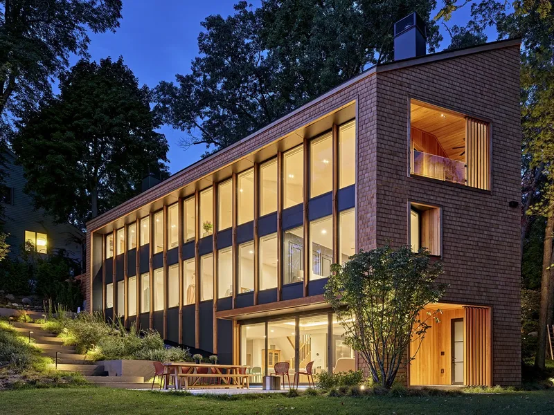 Three-story cedar shingle facade with ribbon windows illuminated at twilight beside a lawn and plantings