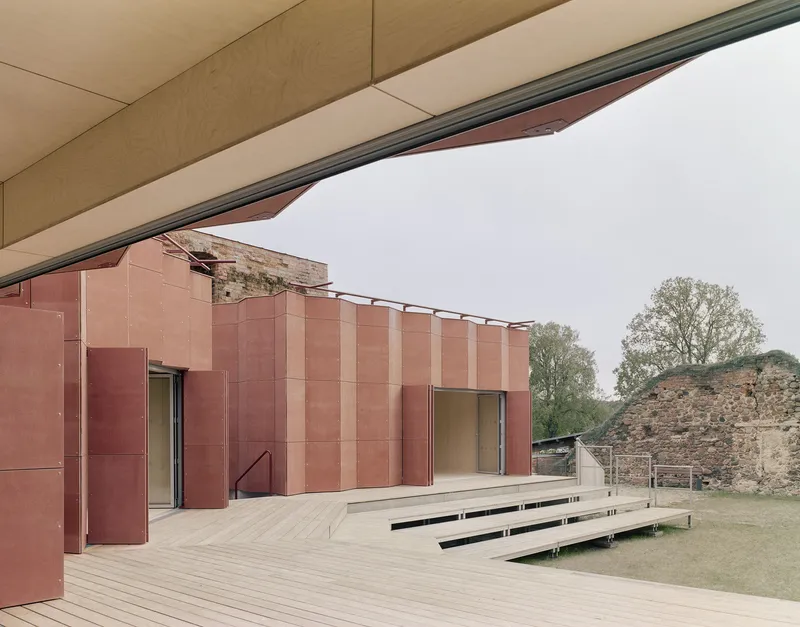 View from beneath a cantilevered plywood ceiling toward copper facade and timber deck with benches