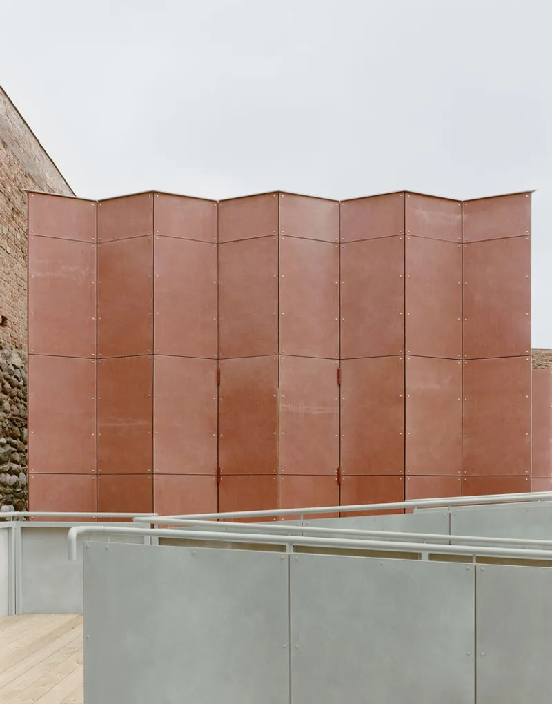 Folded copper panel facade with visible fasteners above a concrete balustrade and timber handrail
