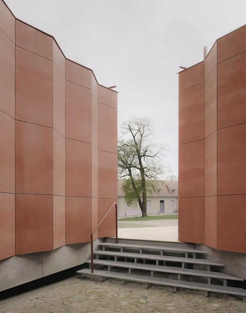 Narrow passage between copper-paneled walls framing a bare tree and distant building on overcast day