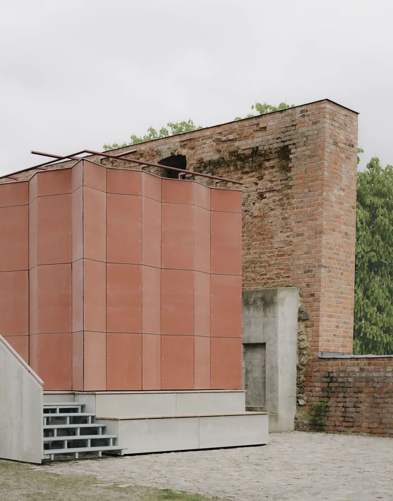 Copper panel cladding meeting deteriorated brick ruin wall under an overcast sky with trees behind