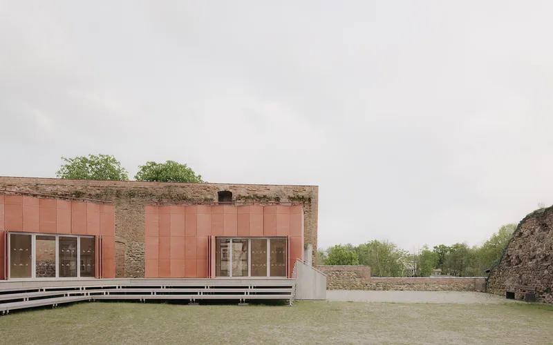 Gravel courtyard with tiered seating facing copper-clad pavilion within crumbling brick fortress walls