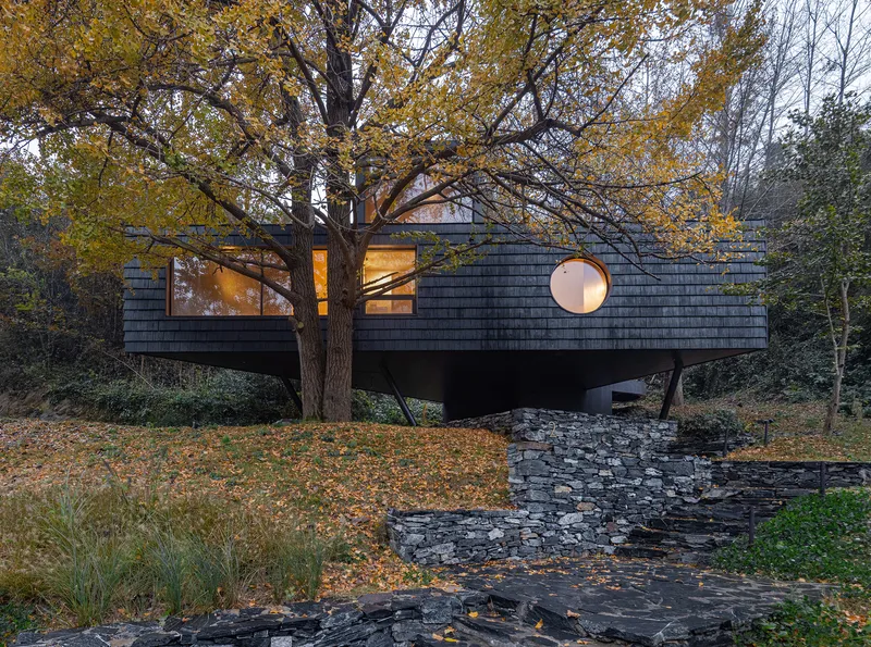 Elevated facade with circular porthole window and dark shingle cladding viewed through autumn trees