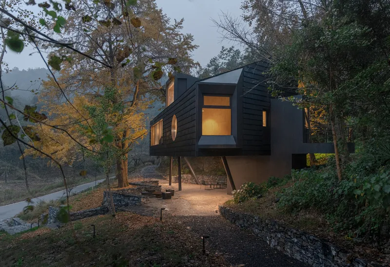 Dark timber-clad volume elevated on slender columns above a gravel clearing at dusk