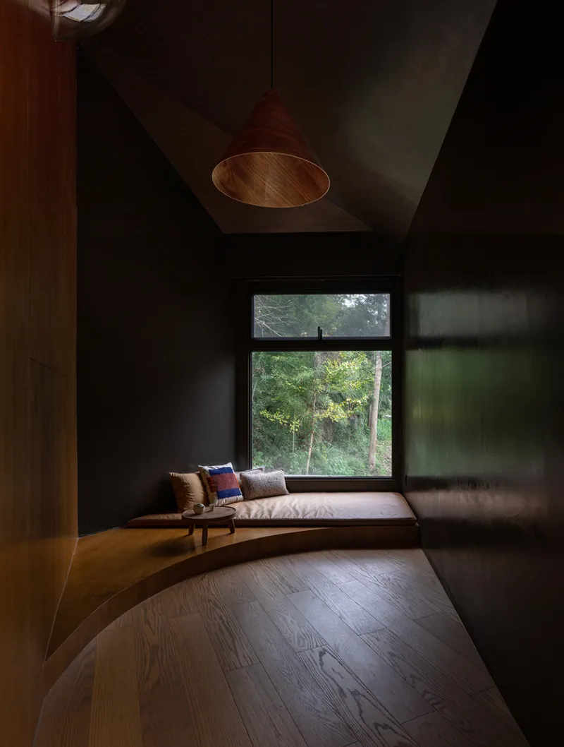 Reading alcove with cushioned bench beneath horizontal window overlooking forest in dim afternoon light