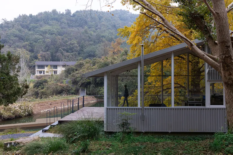 Corrugated metal pavilion with sloped roof and glazed walls nestled among deciduous trees