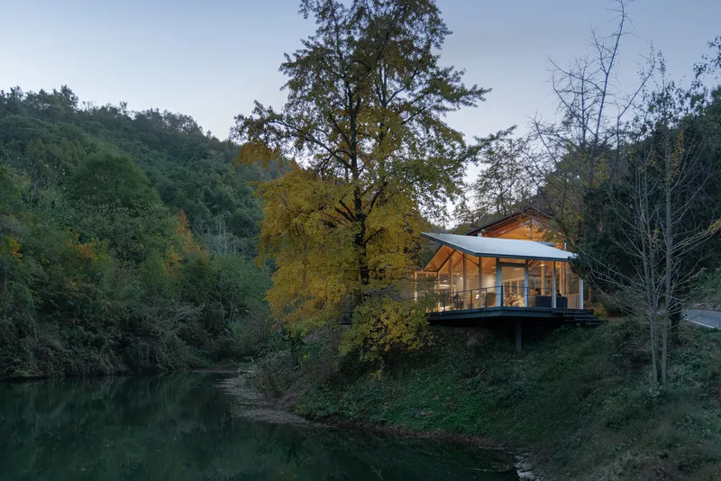 Glass-fronted volume on stilts above a pond surrounded by autumn foliage at dusk