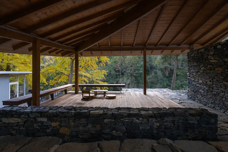 Sheltered terrace with exposed timber rafters and dry-stacked stone walls facing trees