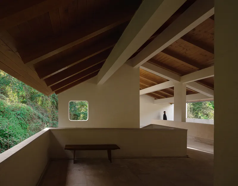 Open-air terrace with exposed timber beams, square oculus window and a solitary bench overlooking the landscape