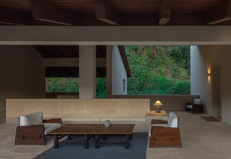 Open living space with exposed timber ceiling beams and large horizontal window framing a green hillside