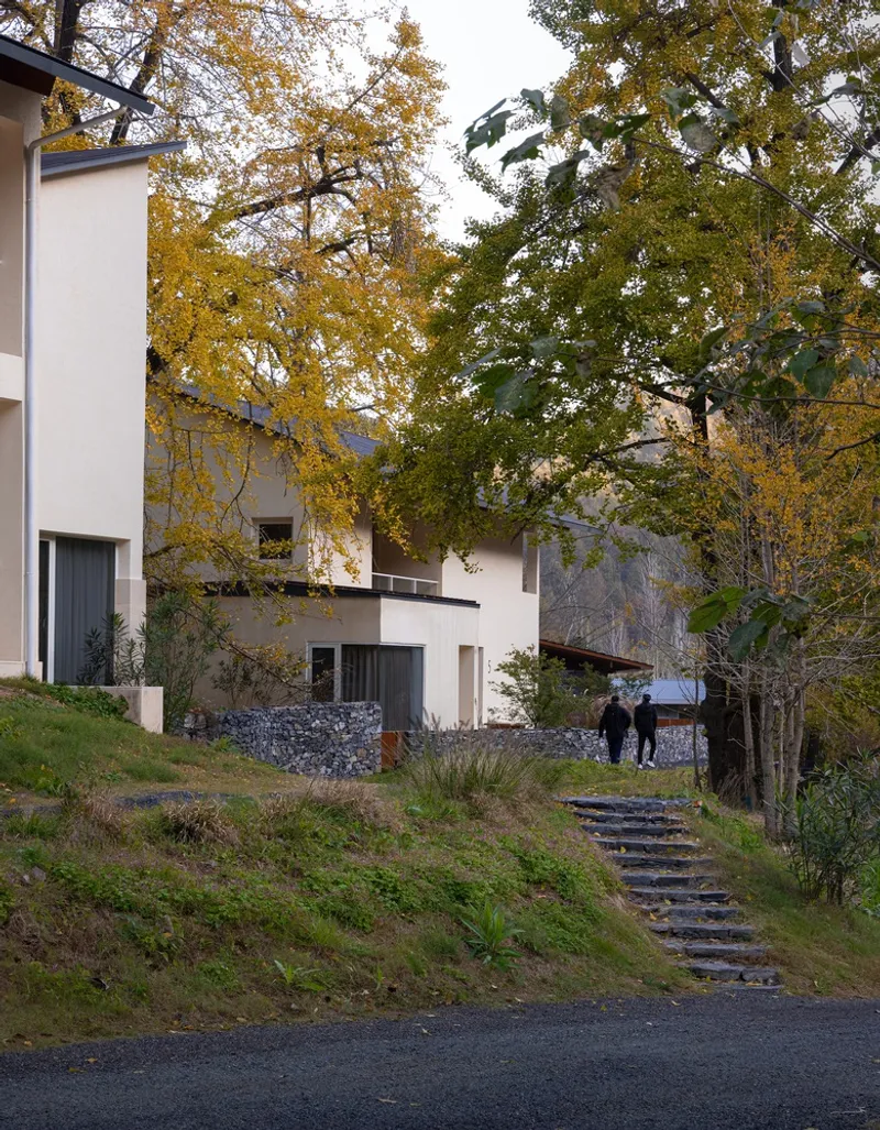 Exterior view showing white plaster volumes and stepped stone path with two figures under autumn canopy