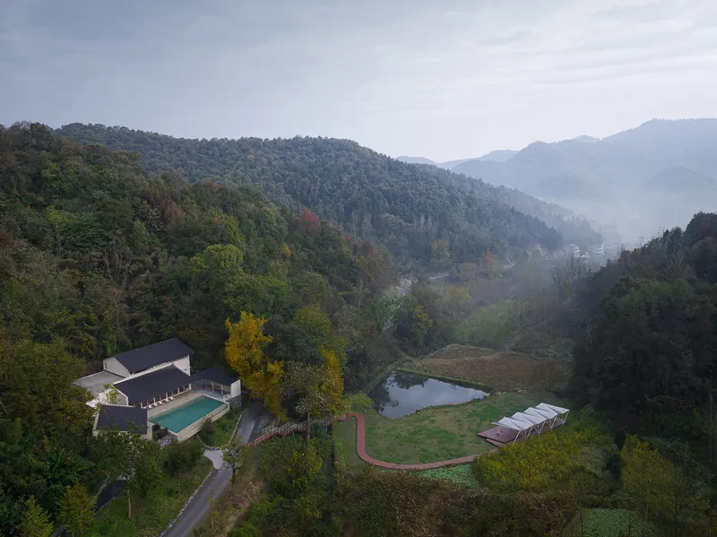 Aerial view of compound with pool and small pond nestled in forested mountain valley at dusk