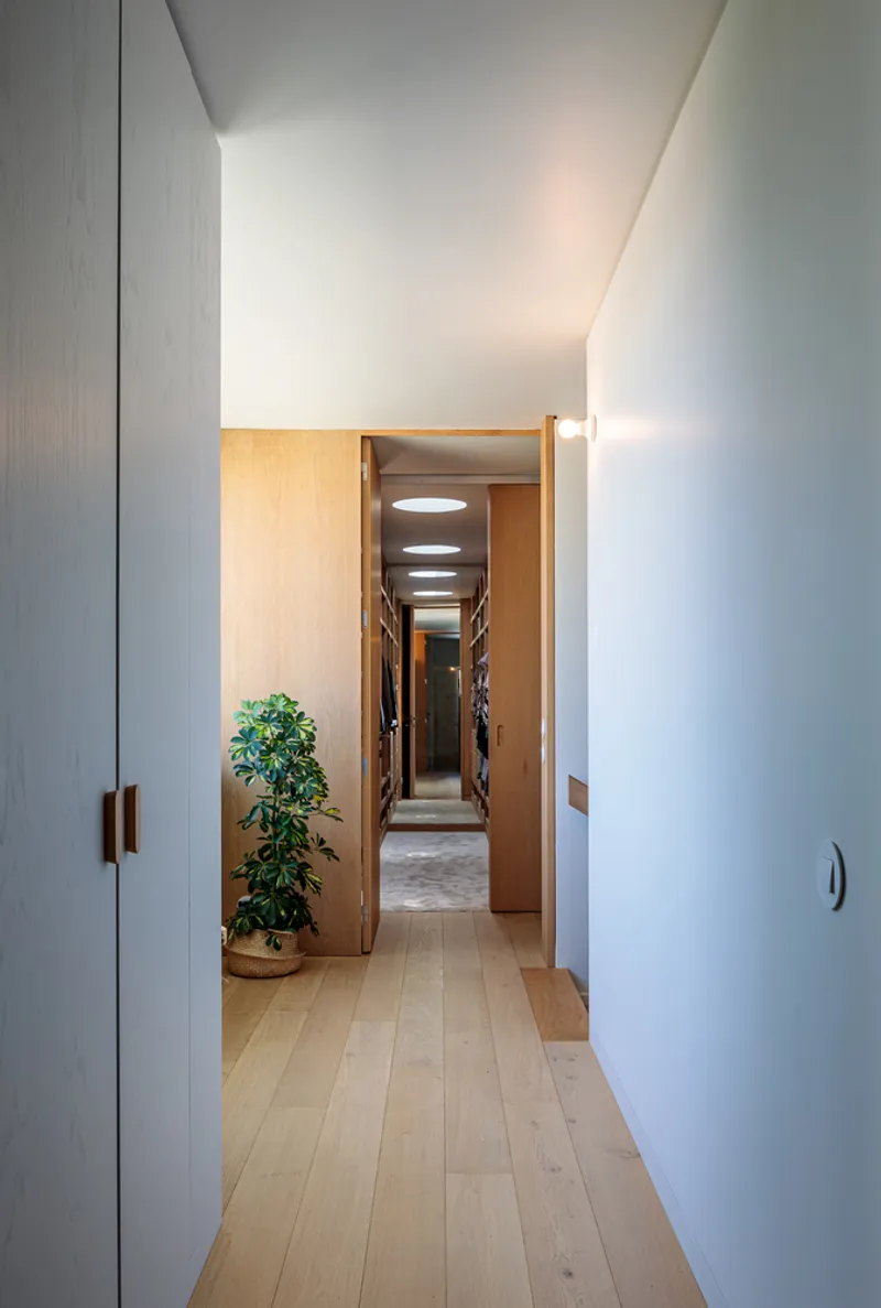 Corridor with light timber flooring and white walls leading to illuminated passage beyond