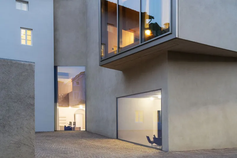 Cantilevered glass bay above a ground-level entrance in a cobbled courtyard at dusk