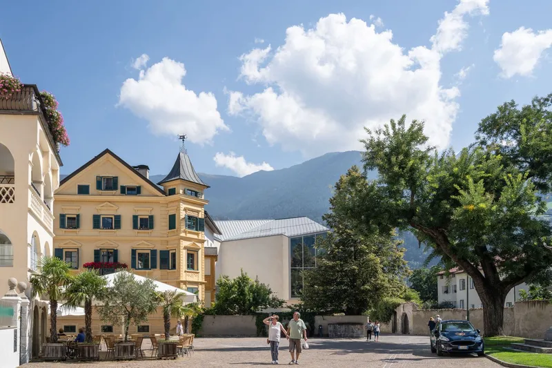 Street view showing the metal-roofed volume among traditional buildings with pedestrians and mountain backdrop