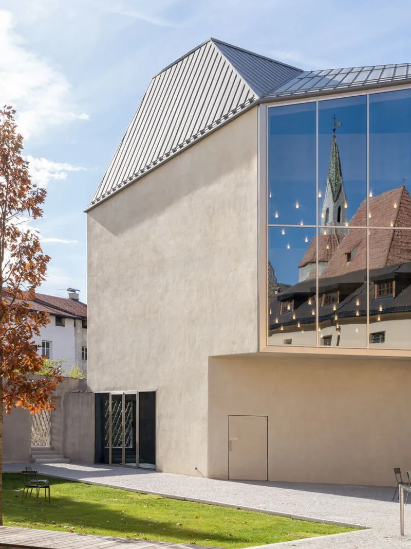 Street view of the beige plaster facade with standing-seam metal roof and tall glass curtain wall reflecting neighboring buildings