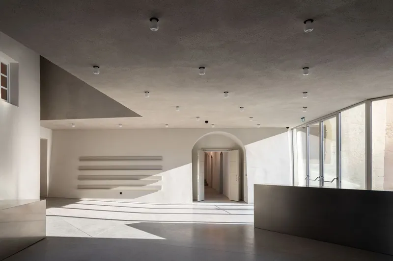 Interior gallery space with curved white staircase, arched doorway, and sunlight streaming across the polished floor
