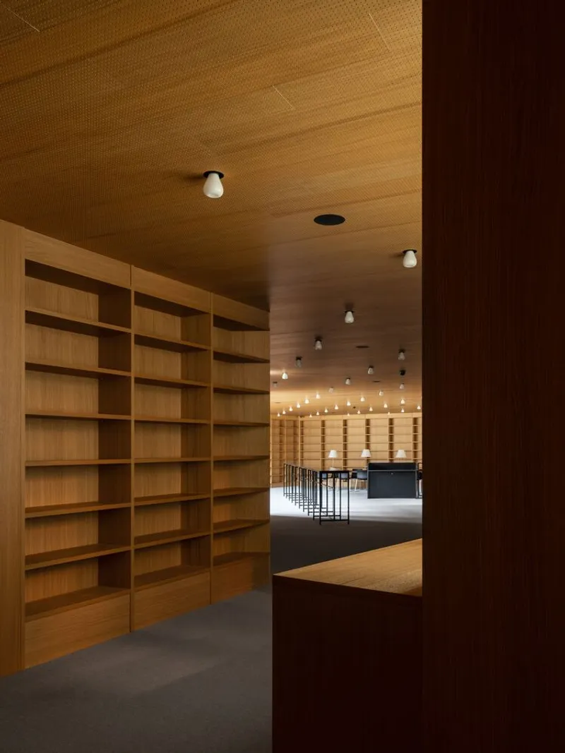 Reading room with floor-to-ceiling timber bookshelves, wood-paneled ceiling, and view through to illuminated storage beyond