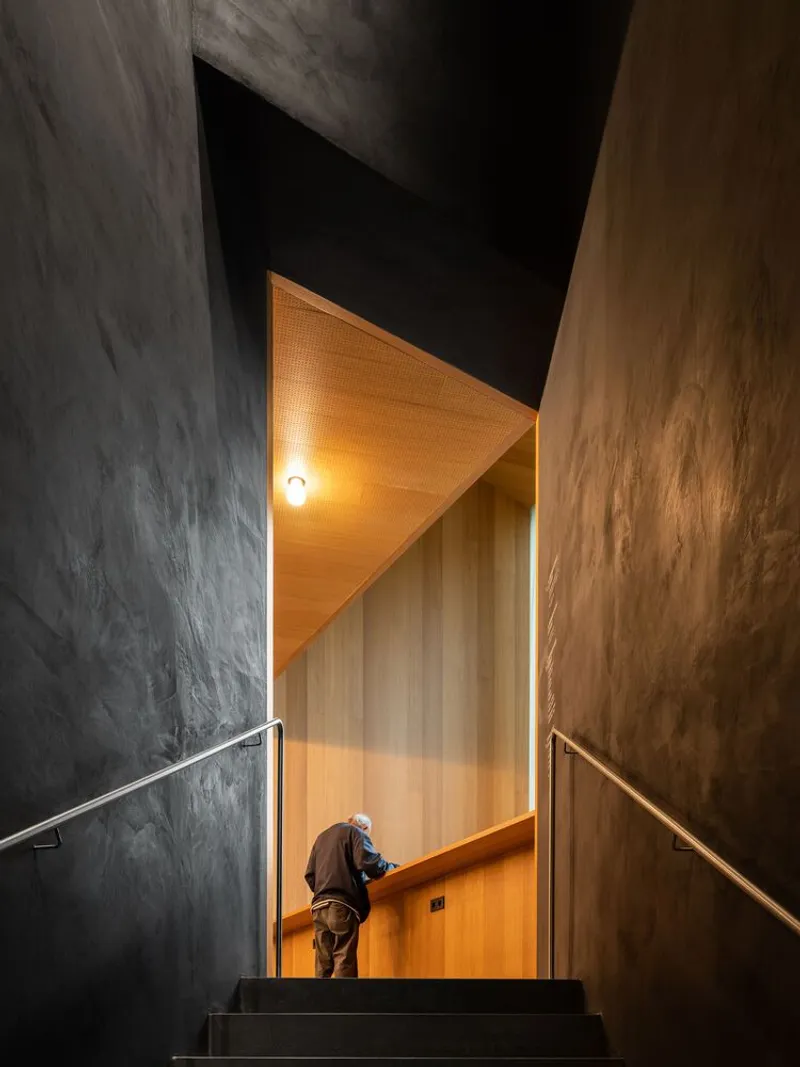 Triangular timber-lined passageway with a visitor ascending the steel stair beneath dark concrete walls