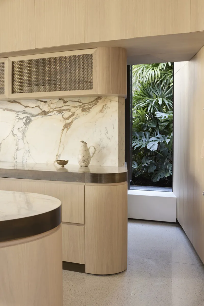 Kitchen island with veined marble backsplash and timber cabinetry beside a narrow window framing tropical foliage