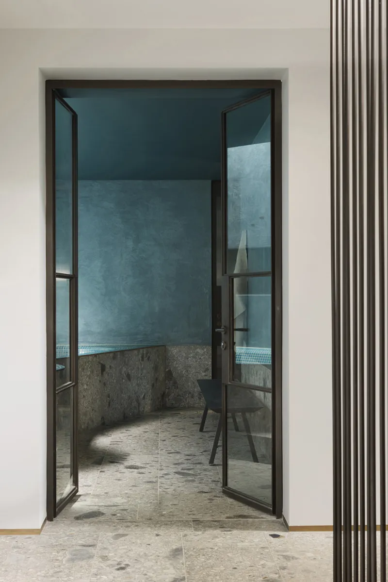 Glazed double door opening from white corridor onto blue plastered bathing room with stone floor